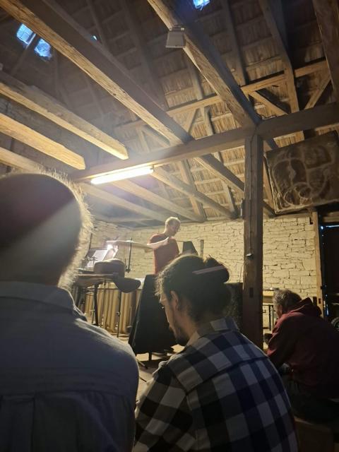 Performer u činelu v kamenné stodole, v popředí publikum